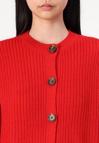 Femme portant un cardigan tricoté rouge vif avec de gros boutons ronds marron, montrant le haut de la poitrine et le bas du visage.