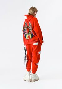 Persona con sudadera con capucha roja y pantalones con diseño de alas coloridas en la espalda, de pie y mirando por encima del hombro, con botas blancas visibles.