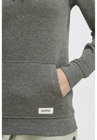 Sudadera gris de tejido suave con un bolsillo frontal, que presenta una pequeña etiqueta blanca con el nombre de la marca "oxmo" en el dobladillo.