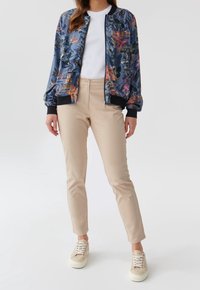 Bavlněná bomber bunda s květinovým vzorem v modré barvě s růžovými a zelenými akcenty, spárovaná s béžovými přiléhavými kalhotami a světlými teniskami. Texturovaná tkanina, na zip.