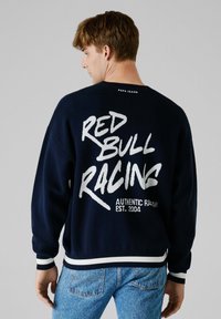 Granatowa bluza z dużym białym nadrukiem "RED BULL RACING" z tyłu, z kontrastowymi białymi i granatowymi ściągaczami przy mankietach i dole.