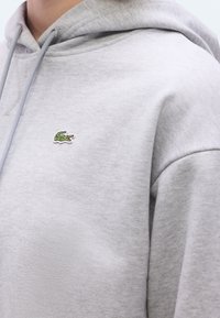 Pehmeast kangast heleda halli fliisiga kapuutsiga pusaga. Sellel on kängurutasku ja vasakul rinnal väike roheline brodeeritud Lacoste'i logo.