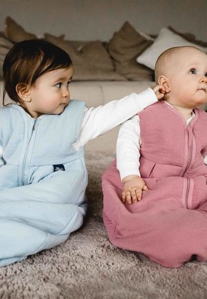 Two seated infants wear sleep sacks. One is light blue, the other pink, both have zippers and soft, textured fabric.