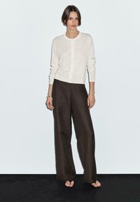 Una donna è in piedi contro uno sfondo chiaro, indossando un cardigan chiaro e pantaloni larghi scuri, con capelli corti e pochi accessori.