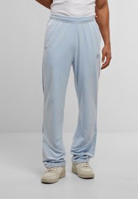 Pantaloni della tuta di velluto morbido in azzurro chiaro con vita elasticizzata, passanti laterali e un logo sull'anca sinistra. Abbinati a scarpe da ginnastica color crema.