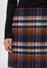 Jupe en laine à carreaux comprenant des couleurs bleu marine, marron, beige et rouge avec une texture douce. Une main placée près de l'ourlet montre les couches de tissu.