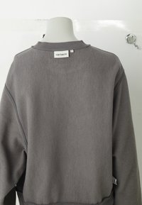 Harmaa fleece-huppari, jossa on pyöreä kaula-aukko, ribattu hihansuu ja helma, sauma yksityiskohtia sekä pieni brändilippu takana.