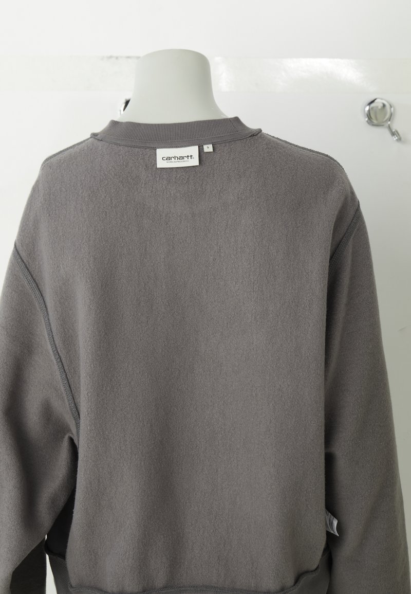 Grå fleece sweatshirt med rund halsringning, ribbade ärmslut och nederkant, med sömnadsdetaljer och en liten varumärkesetikett på baksidan.