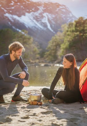 Twee mensen in donkere buitenkleding zitten op de zandgrond naast een rivier, koken met een zilveren pan. Een feloranje tent is op de achtergrond te zien.