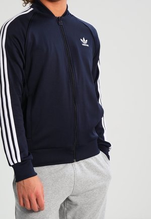 Osoba ubrana w granatową kurtkę sportową Adidas z białymi paskami na rękawach i jasnoszare spodnie dresowe, stojąca z jedną ręką w kieszeni.