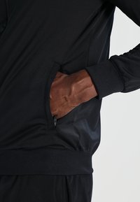 Veste de sport noire avec une poche latérale zippée, des poignets côtelés et une texture lisse. Gros plan d'une main insérée dans la poche.