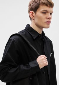 Μαύρο πουκάμισο με κουμπιά και γιακά, με διακριτική κεντημένη λεπτομέρεια. Λείο ύφασμα, εφαρμοστή γραμμή, φορεμένο με μαύρο λουρί τσάντας στον ώμο.