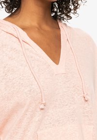 Pullover con cappuccio rosa chiaro realizzato in tessuto morbido, con scollatura a V, dettagli con cordini e tasca frontale. Design casual con texture.