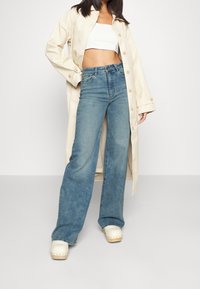 Vaqueros de pierna ancha en denim azul, combinados con un top corto blanco y un abrigo largo beige claro. El conjunto incluye zuecos de madera.