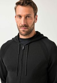 Schwarze Kapuzenjacke mit Reißverschluss und strukturiertem Design, ausgestattet mit einer Kordelzugkapuze und gerippten Details an den Schultern und Seiten.