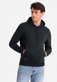 Felpa nera con cappuccio e tasca frontale a marsupio, con coulisse sul cappuccio, polsini a costine e un sottile patch del logo sull'orlo. Texture morbida, design casual.