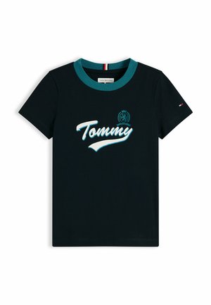 Uśmiechnięte dziecko z kręconymi włosami stoi z rękami za plecami, ubrane w granatowy t-shirt "Tommy", luźne niebieskie dżinsy i białe trampki.