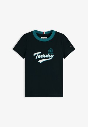 Dijete s osmijehom i kovrčavom kosom stoji s rukama iza leđa, nosi tamnoplavu majicu "Tommy", široke plave traperice i bijele tenisice.