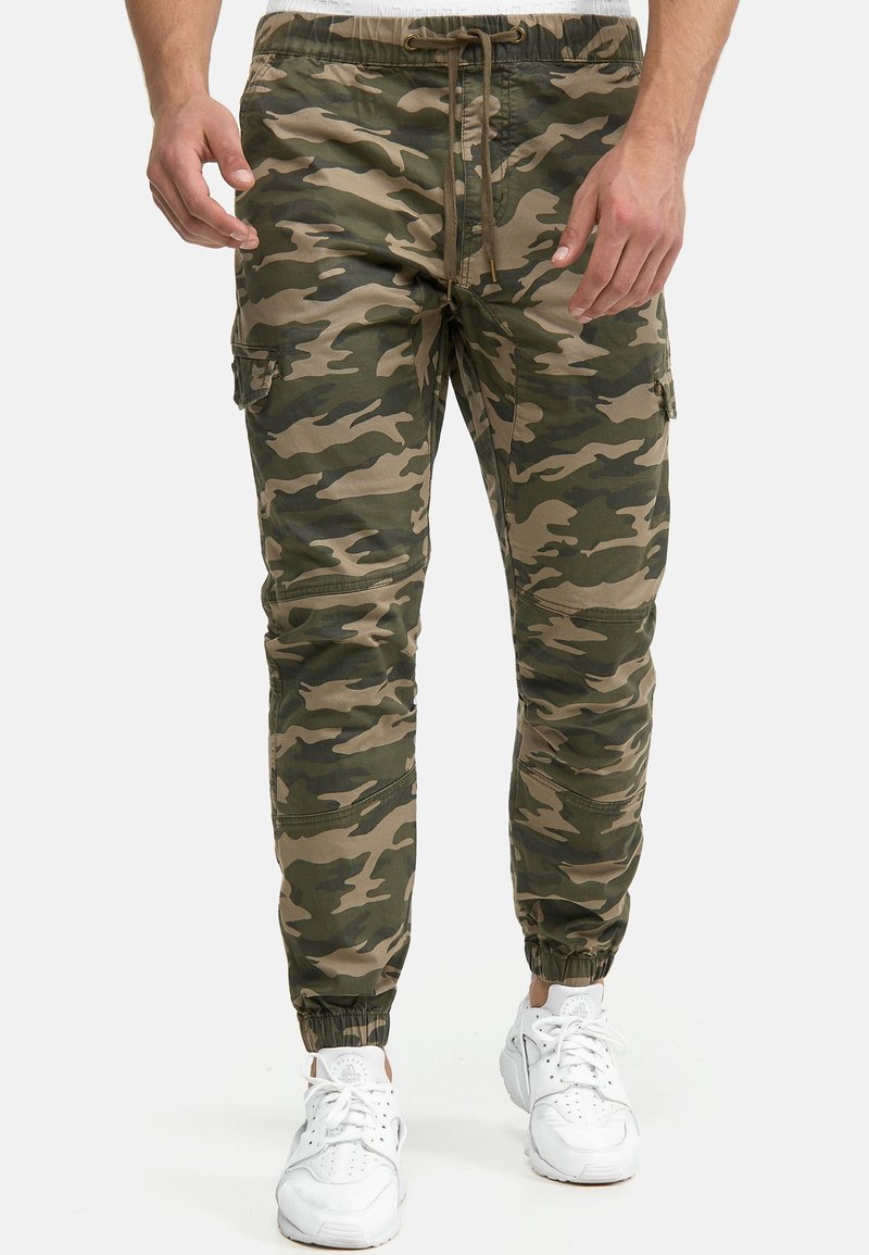 Homme portant un pantalon cargo camouflage vert et beige avec taille à cordon et poignets élastiques, associé à des baskets blanches.