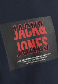 Námořnické tričko s velkým grafickým potiskem s nápisem "JACK & JONES" v červené a černé barvě, s dalším textem dole v bílé. Hladký bavlněný materiál.