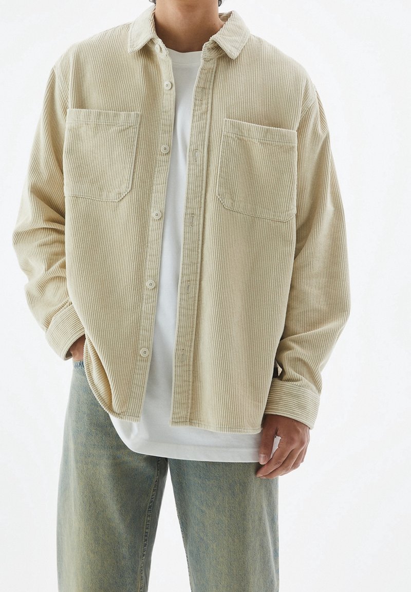 Beige Waffle-Shirtjacke mit Knopfverschluss, zwei Brusttaschen und entspannter Passform, über einem weißen T-Shirt und hellblauen Jeans getragen.