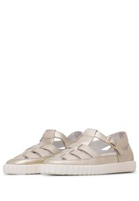 Metallisch goldene flache Sandalen mit einem geflochtenen Design, offenen Seiten und einem gewellten Knöchelriemen. Die Sohle ist weiß und rund, mit strukturierten Rillen.