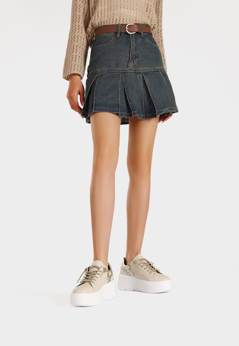 Gonna plissettata in denim blu scuro con vita aderente e cintura marrone. Indossata con sneakers platform beige caratterizzate da una texture liscia e dettagli.