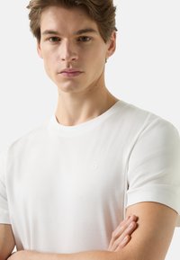 Biały bawełniany t-shirt z krótkimi rękawami, okrągłym dekoltem i małym, wytłaczanym logo na piersi. Gładka faktura, dopasowany krój.