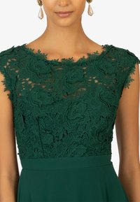 Grünes Spitzenkleid mit Blumenmustern, einem transparenten Ausschnitt und einem glatten Stoffrock. Es hat ein figurbetontes Oberteil und kurze Ärmel.