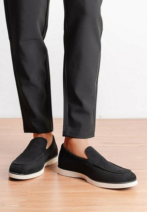 Personne portant des chaussures noires sans lacets avec des semelles blanches et un pantalon noir ajusté, debout sur un sol en bois.