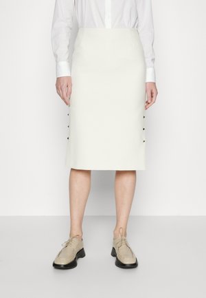 Kvinde bærer hvid blyantsnederdel til knæet, hvid skjorte med knapper og beige snøresko, står mod en ensfarvet hvid baggrund.