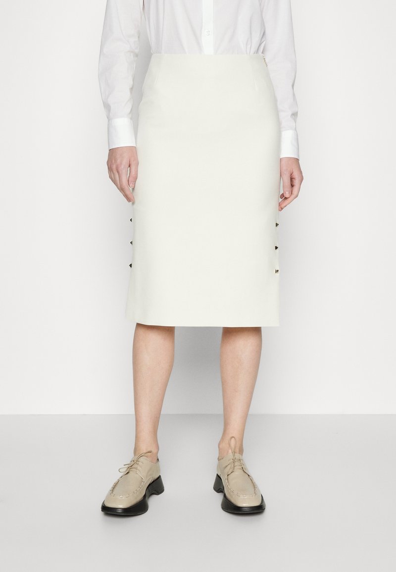 Femme porte une jupe crayon blanche mi-genou, une chemise blanche boutonnée et des chaussures beiges à lacets, debout sur un fond blanc uni.