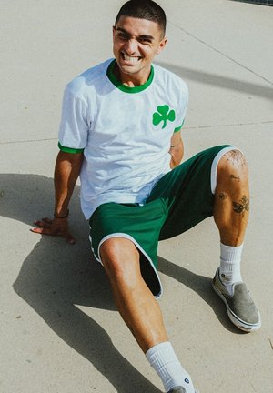 Hvid sports t-shirt med et grønt kløverlogo og grøn krave, parret med grønne træningsshorts, hvide sokker og grå slip-on sko.