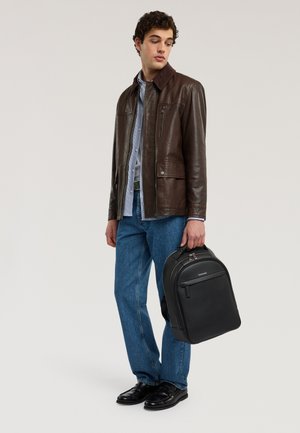 Junger Mann mit brauner Lederjacke, blauer Jeans und schwarzen Schuhen, hält einen schwarzen Rucksack am oberen Griff und schaut nach unten zur Seite.