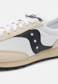 Hvid og beige sportsko med tekstureret ruskind og glat stof, med et sort accentlogo og cirkulære perforeringer på siden.