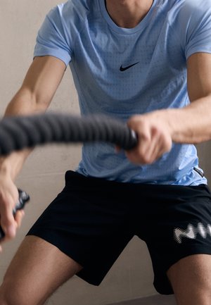 Man in blauw Nike-shirt en zwarte shorts die traint met battle ropes, focus op armen en torso.