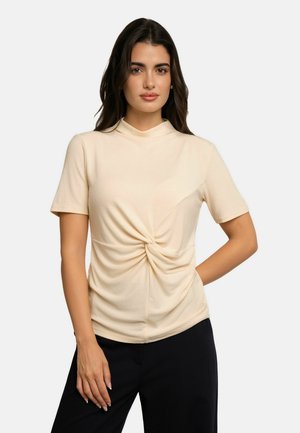 Jeune femme aux longs cheveux foncés portant un haut crème à manches courtes avec un détail torsadé à l'avant et un pantalon noir, debout devant un fond uni.