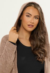 Cárdigan de canalé marrón claro con capucha, que presenta mangas largas y un acabado texturizado, combinado con una blusa negra ajustada por debajo.