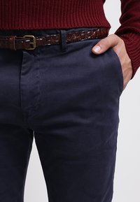 Mann trägt marineblaue Hosen und einen braunen Ledergürtel mit einer Messingschnalle, Hand in der Tasche ruhend, und einen roten gestrickten Pullover.