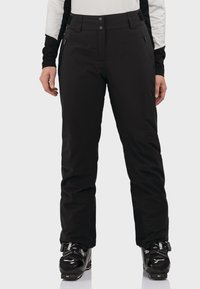 Schwarze Skihosen mit passgenauer Passform, ausgestattet mit Reißverschlusstaschen und Gürtelschlaufen. Kombiniert mit schwarzen Skischuhen für den Wintersport.