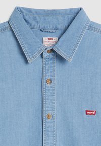 Camicia di jeans blu chiaro con colletto a punta, bottoni marroni e patch logo rosso Levi's sul lato sinistro del petto. Texture morbida e design casual.