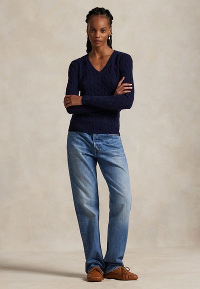 Woman standing with arms crossed wearing a navy cable-knit V-neck sweater, loose blue jeans, and brown moccasin shoes against a plain background.