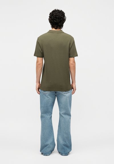 Homme aux cheveux bouclés portant un polo vert olive à manches courtes et un jean bleu clair à jambes larges, debout de dos devant un fond blanc.