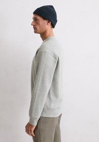 Grijze sweatshirt met een ronde hals en ribgebreide manchetten, gedragen met olijfgroene broeken. Een donkerblauwe ribgebreide beanie maakt de look af.