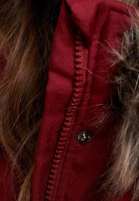 Röd tygjacka med pälskantad huva, som har en robust dragkedja och tryckknapp. Närbild visar sömnadsdetaljer och textur.