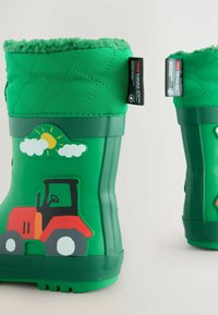 Botas de goma verdes con parte superior acolchada, que presentan un diseño de tractor rojo, sol y nubes. Suelas texturizadas para agarre, forro interior suave.