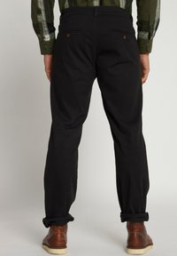 Pantaloni neri con orli arrotolati, dotati di due tasche posteriori con bottoni marroni, realizzati in un tessuto morbido. Indossati con scarpe di pelle marroni.