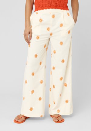 Pantalon large couleur crème avec des imprimés de soleil orange, porté avec des tongs orange et un top en maille orange sans manches.