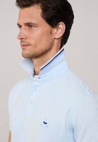 Polo bleu ciel avec un col relevé, deux boutons et un logo sur la poitrine. Le col présente des rayures bleu marine et blanches. Texture du tissu douce.