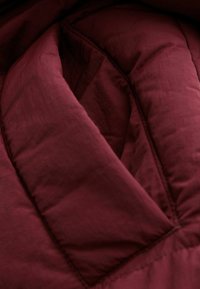 Bordeauxrote Steppdaunenjacke mit gestepptem Design, zeichnet sich durch eine glatte Textur und sichtbare Nahtlinien aus. Weiches, isoliertes Material.